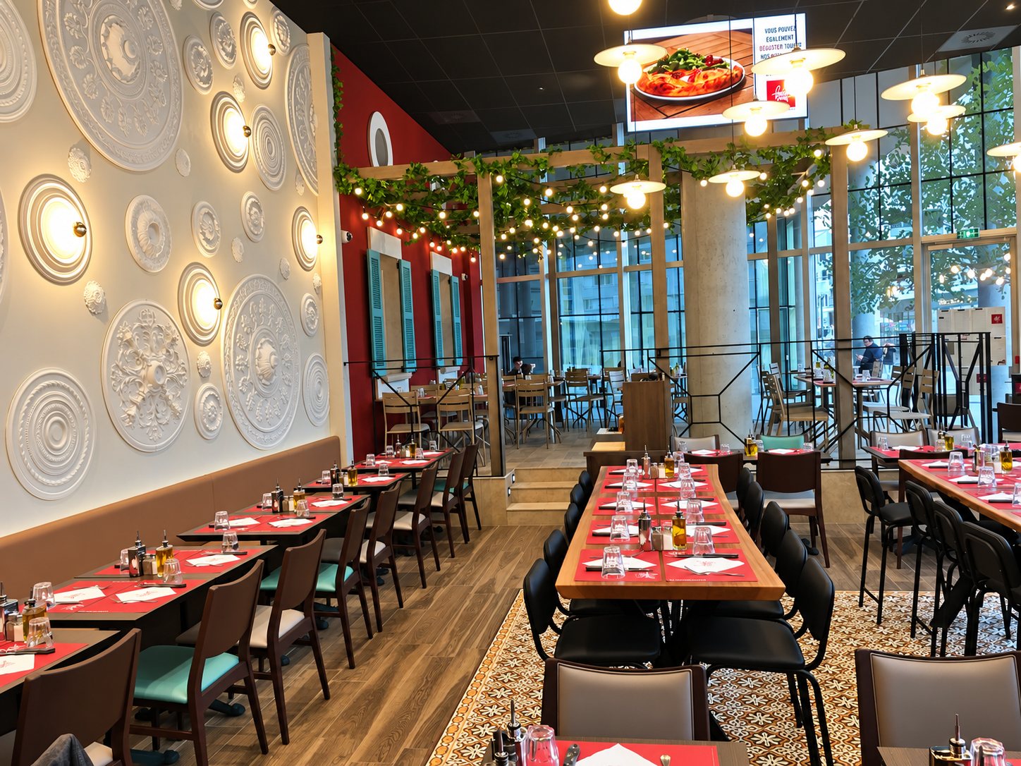 Intérieur d'un restaurant avec de longues tables dressées de sets rouges, un mur blanc orné de rosaces lumineuses et des guirlandes lumineuses au plafond.