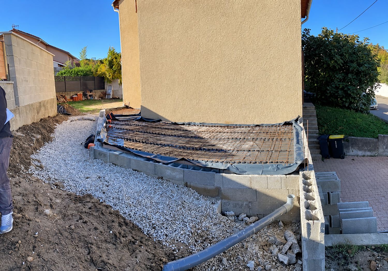 Terrasse en béton en cours de réalisation contre une maison, avec gravier, parpaings et treillis métallique.