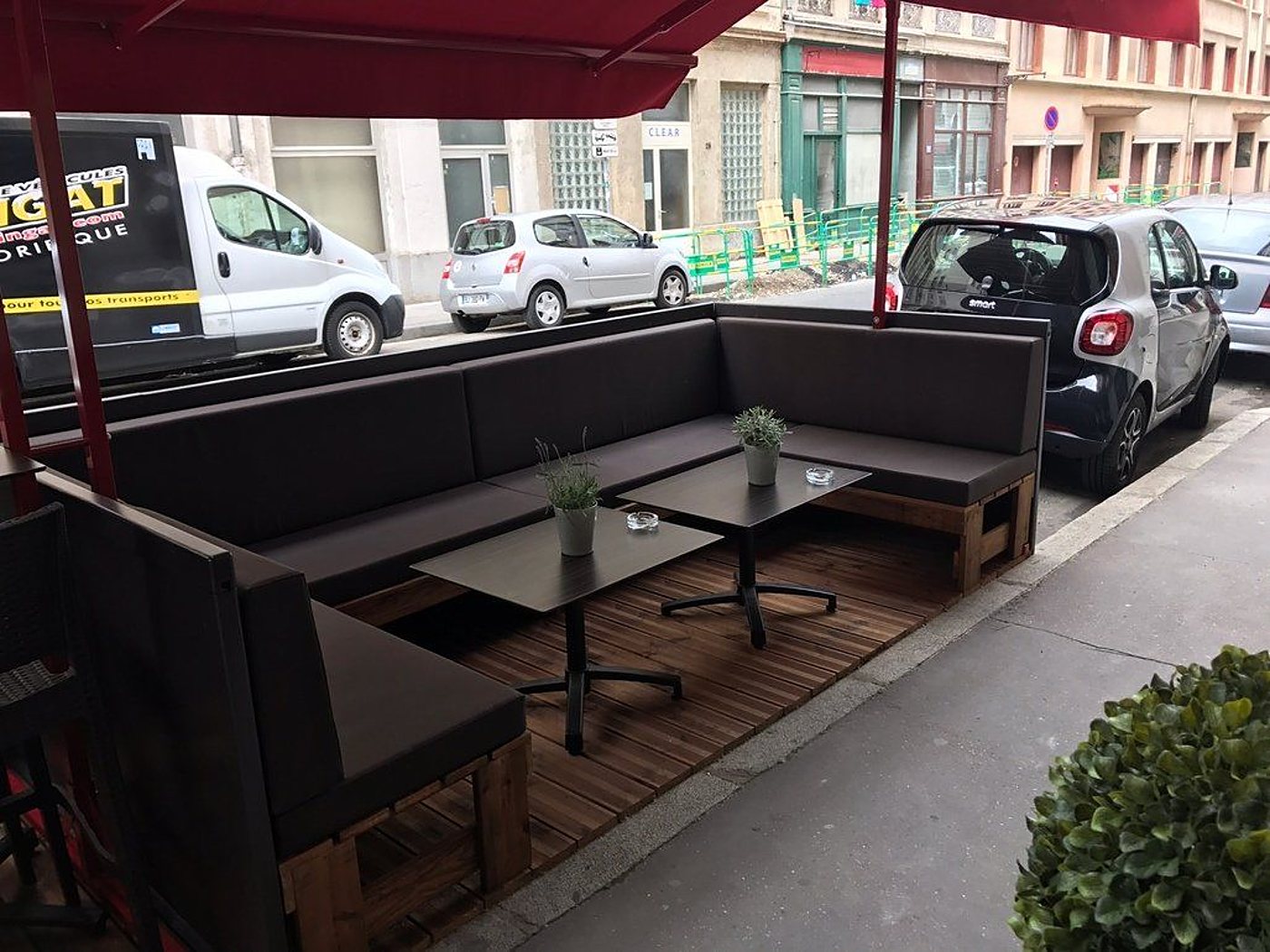 Terrasse extérieure sous un store rouge avec une grande banquette d'angle brune, deux tables basses et un plancher en bois, au bord d'une rue avec des voitures garées.