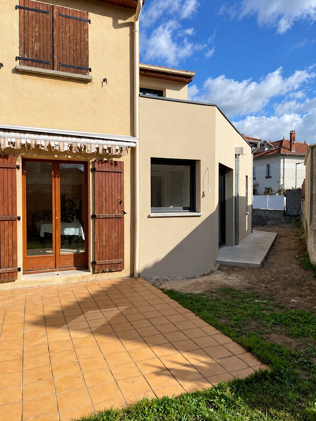 Terrasse carrelée et pelouse devant une maison beige à étage avec une extension récente à toit plat.