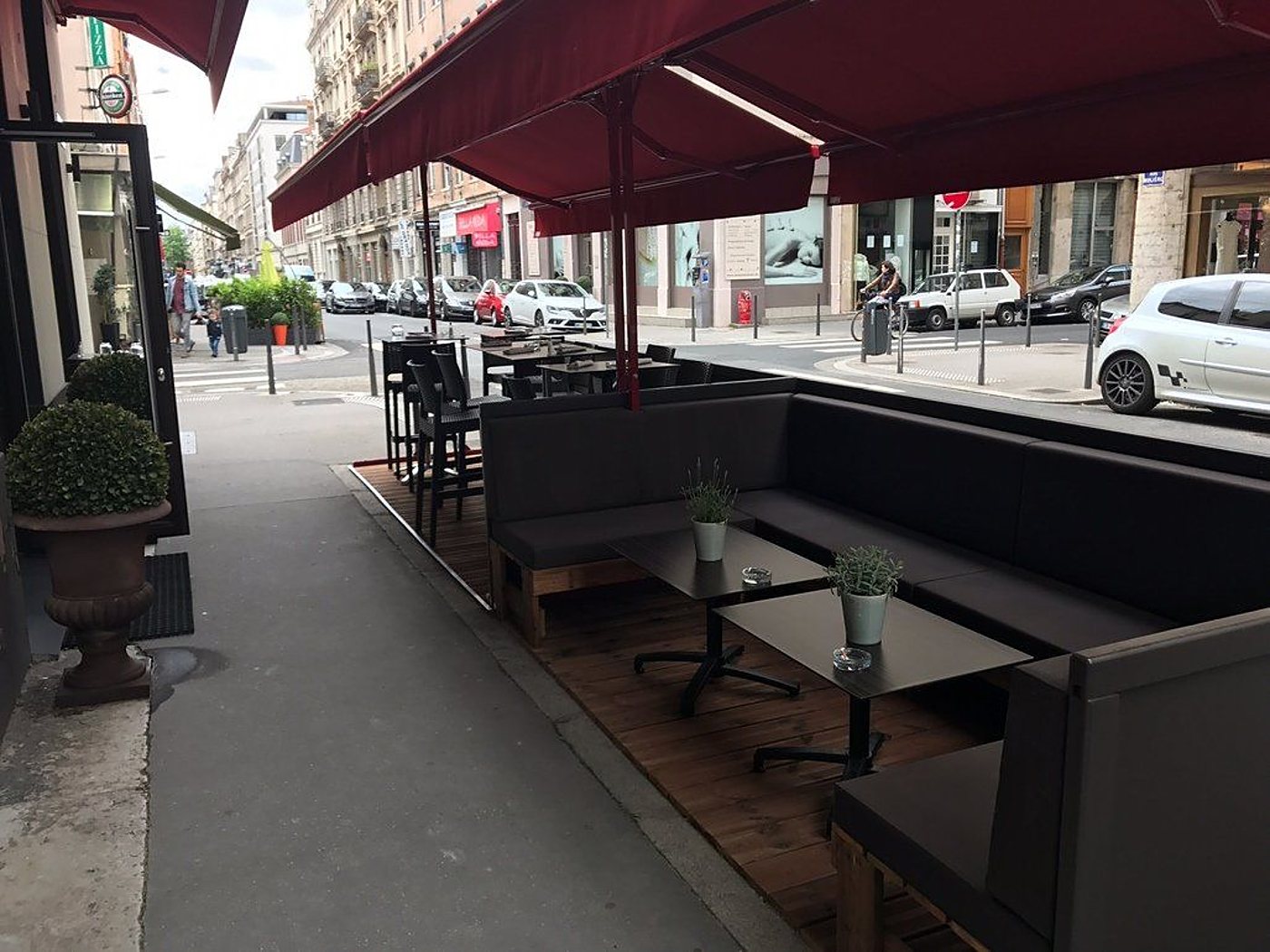 Terrasse de café sous un auvent bordeaux, avec banquettes sombres, tables basses et plantes en pot, donnant sur une rue urbaine.