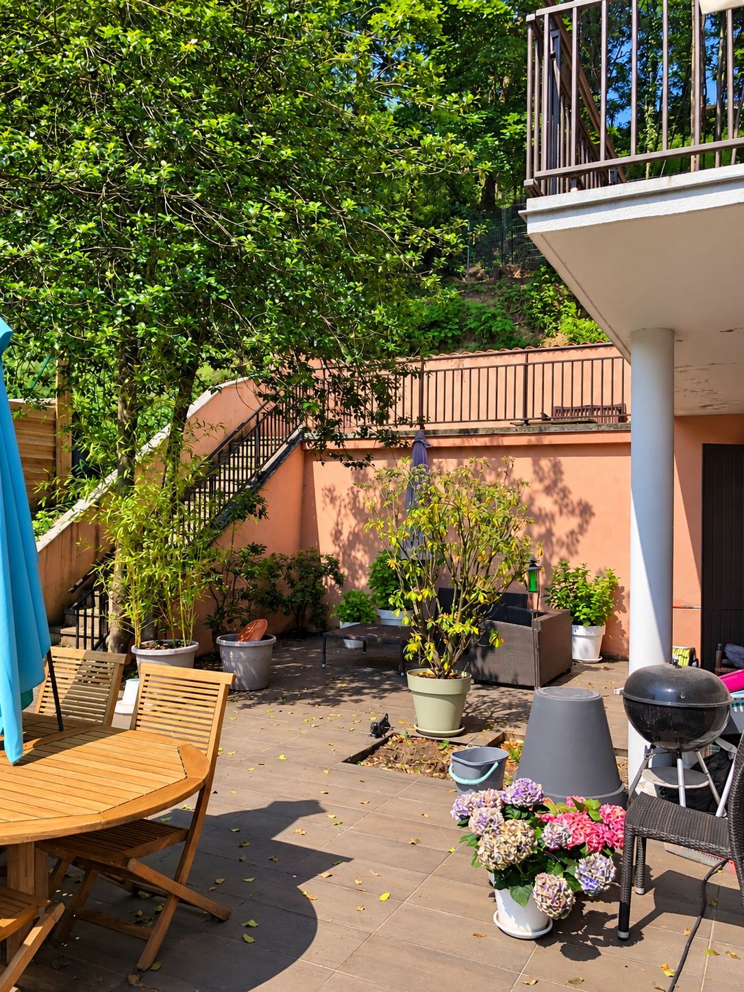 Une terrasse carrelée et ensoleillée avec une table et des chaises en bois, des hortensias en pots, un barbecue et un escalier en arrière-plan.