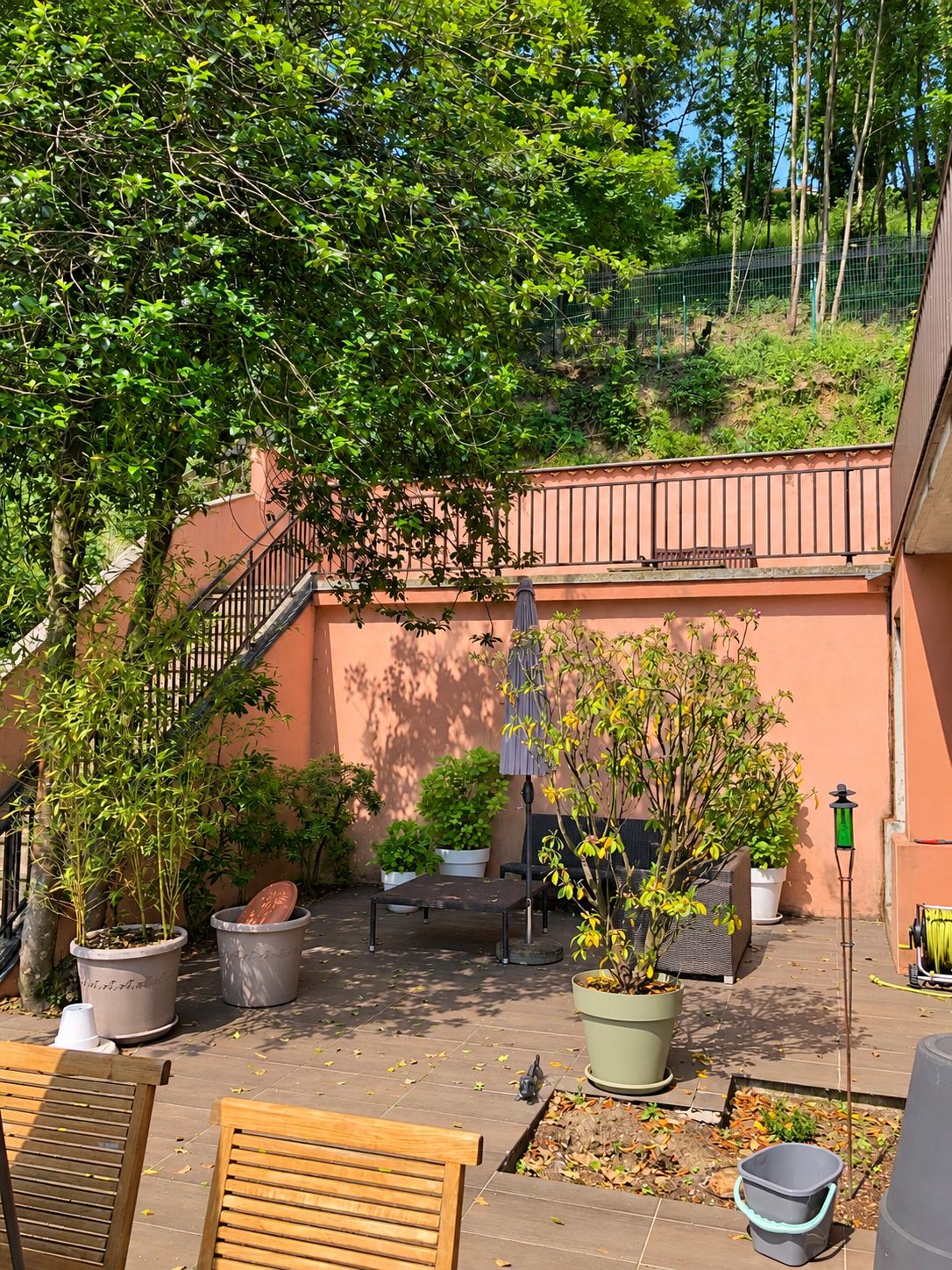 Une terrasse carrelée avec des plantes en pots, un parasol fermé et des chaises en bois devant un mur rose et un escalier, au pied d'un talus boisé.