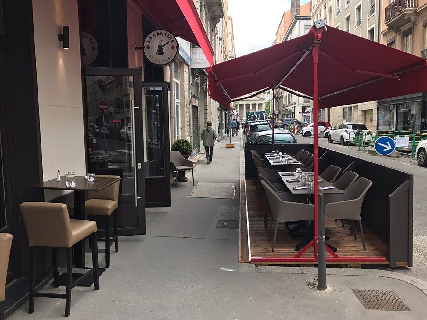 La terrasse du restaurant La Cantine, avec tables dressées et fauteuils gris sous un grand parasol rouge, borde une rue en ville.