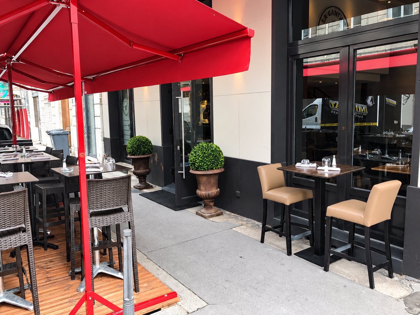 Terrasse d'un restaurant sous un store rouge, avec tables hautes, chaises beiges et deux buis en vasques devant l'entrée vitrée.