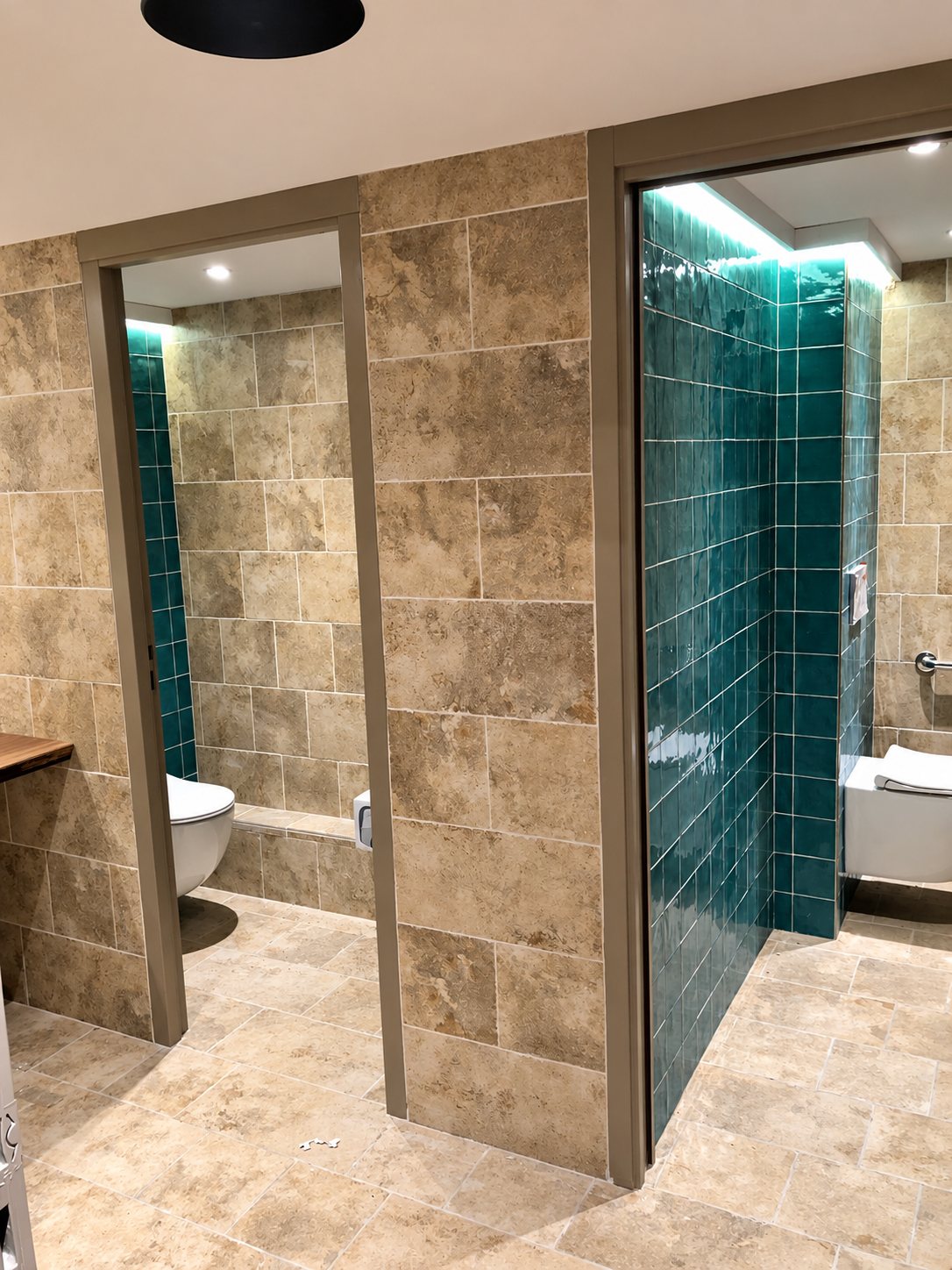 Deux cabines de toilettes aux murs en carrelage beige effet pierre, avec un fond de faïence verte émaillée et des WC suspendus blancs.