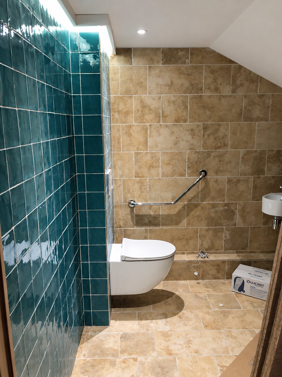 Des toilettes rénovées avec un WC suspendu blanc, une barre d'appui chromée, un mur en carreaux verts brillants et un sol et des murs en pierre beige.
