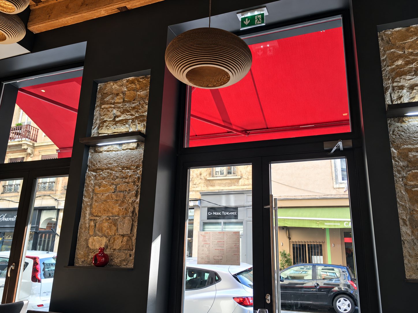 Vue à travers une vitrine à store rouge sur une rue avec des voitures garées.
